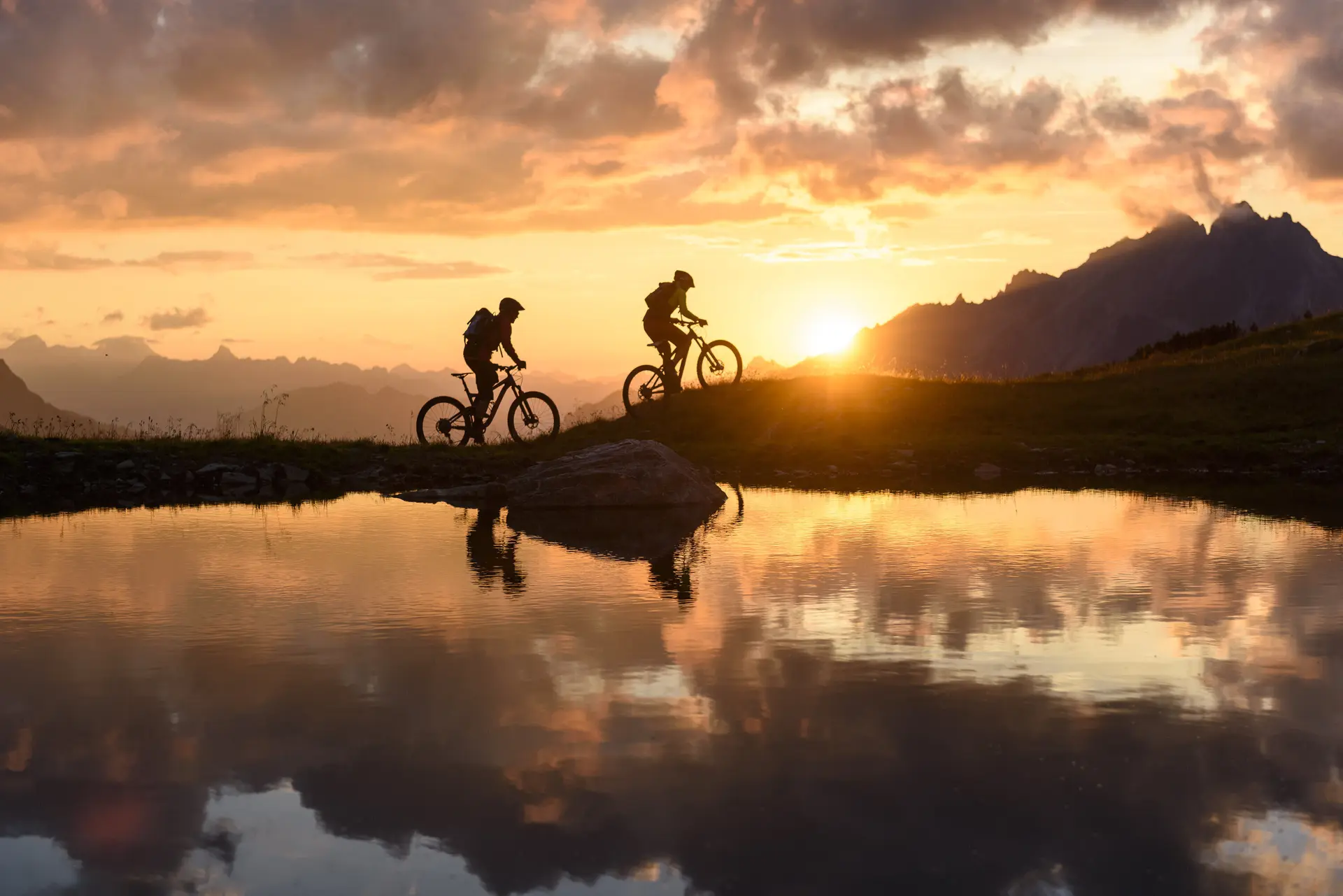 Zwei Personen fahren im Sonnenuntergang mit ihren Mountainbikes vor einer Bergkulisse. | © DAV/Wolfgang Ehn